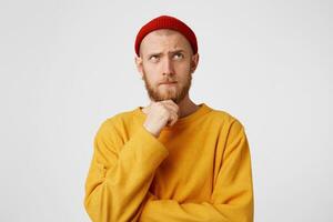 Close up photo of thoughtful man looking aside isolated over white background wall. Waiting for inspiration. Thinking young bearded male in red hat holding fist on chin