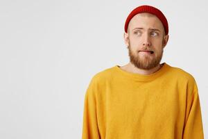 Close up of young guy with a red beard, wearing a red hat, cowardly, cautiously pursing his lip, glances left at the empty space for copy, isolated on white background photo