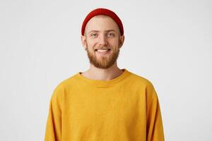 Shot of happy bearded young man, looks with joyful expression, has friendly smile, wears yellow sweater and red hat, isolated over white studio wall. photo