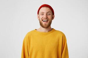 Indoor shot of bearded young cute man, who is amused by his friends, laughs joyfully as hears funny joke, wears red hat and sweater, poses against white wall. Happiness and positive emotions concept photo