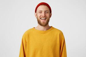 Isolated shot of joyful bearded young cute man laughs joyfully as hears funny joke, wears red hat and sweater, poses against white studio wall. Happiness and positive emotions concept photo