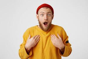 Amazed male keeps mouth opened, hands on breast, being shocked, stares at camera, isolated over white background. Bearded man in red hat man expresses great surprisement photo