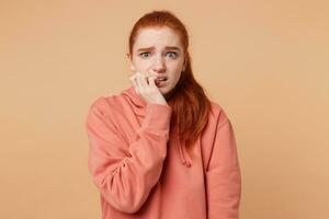 Portrait of a frightened young girl with blue eyes and red hair gathered in a ponytail that looks at the camera with fear,bites her nails, holds hand near the mouth,feels like a victim wants to escape photo