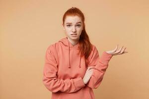 Dissatisfied red-haired girl with a tail dressed in hoodie with someone arguing one hand raised palm up trying to prove something, to defend their opinion, on beige studio background photo