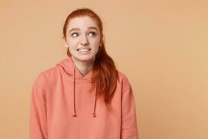 Pretty young red-haired girl with her hair gathered in a ponytail dangerously, with caution uncertainty spies to the right on empty space, feels insecure, isolated on a beige background photo