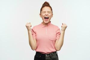 Portrait of happy, celebrating girl with ginger hair bun. Wearing pink t-shirt and black jeans. Clench her fists, keeps eyes closed and scream in excitement. Stand isolated over white background photo