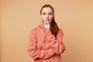 Cute red-haired girl with a hair gathered in ponytail nicely flirts, makes a silence gesture with index finger on her lips. Lets keep a secret, stay quiet, remain a privacy photo