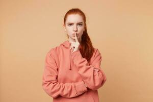 Indoor photo of red-haired serious girl with a tail with beautiful blue eyes, urges to keep silence, to behave quietly, to calm down not to produce noise, isolated on a beige background.