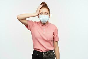 retrato de conmocionado niña con jengibre pelo bollo. vistiendo rosado camiseta y médico, protector cara mascarilla. conmovedor cabeza, recordado algo. acecho a el cámara, aislado terminado blanco antecedentes. foto