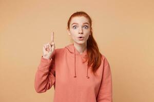Young pretty red haired woman isolated over beige background pointing finger up popping eyes with successful idea. Exited and happy. Number one. photo