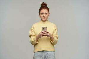 Unhappy looking woman, displeased redhead girl with hair bun. Wearing yellow sweater and holding a smartphone. Have seen something negative. Watching at the camera, isolated over grey background photo