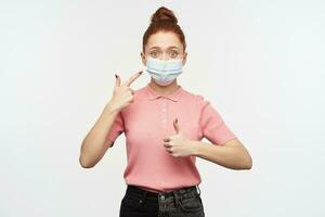 Portrait of girl with ginger hair bun. Wearing pink t-shirt and medical, protective face mask. Pointing at the mask and shows thumb up. Watching at the camera, isolated over white background photo