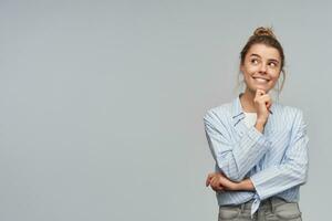Portrait of attractive, dreaming girl with blond hair bun. Wearing blue striped knotted shirt. Touching her chin and watching curious to the left at copy space, isolated over grey background photo