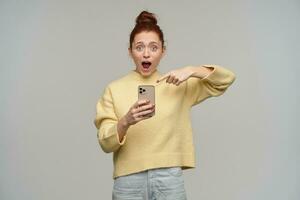 Portrait of attractive, excited girl with ginger hair bun. Wearing yellow sweater and holding a smartphone. Pointing with finger at her phone. Watching at the camera, isolated over grey background photo