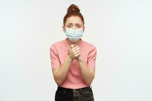 Young, desperate woman with ginger hair bun. Wearing pink t-shirt and protective face mask. Keeps palms together, plead for something. Watching at the camera, isolated over white background photo