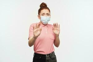 Portrait of horrified, adult girl with ginger hair bun. Wearing pink t-shirt and protective face mask. Pushes arms away, protect herself. Watching at the camera, isolated over white background photo