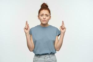 Portrait of attractive, cute girl with ginger hair bun. Wearing blue t-shirt. Biting lip and keeps fingers crossed. Making a wish. Watching at the camera, isolated over white background photo