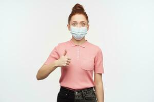 Happy, excited redhead girl with hair bun. Wearing pink t-shirt and medical, protective face mask. Showing thumb up sign, approval. Watching at the camera, isolated over white background. photo