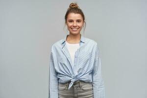 Happy woman, positive blond girl with hair bun. Wearing blue striped knotted shirt. Standing confident, broadly smiling. Emotion concept. Watching at the camera, isolated over grey background photo