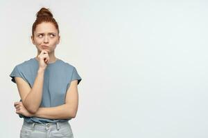 Portrait of attractive, dreaming girl with ginger hair bun. Wearing blue t-shirt. Touching her chin and thinks about something. Watching to the right at copy space, isolated over white background photo