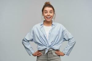 Portrait of excited, positive girl with blond hair bun. Wearing blue striped knotted shirt. Emotion concept. Holds hands on a waist. Watching surprised at the camera, isolated over grey background photo