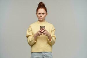 ceñudo pelirrojo niña con pelo bollo y fruncido labios. vistiendo amarillo suéter y participación un teléfono inteligente teniendo pensamientos acerca de contenido. acecho confuso a el cámara, aislado terminado gris antecedentes foto