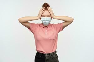 Stressed woman with ginger hair bun. Wearing pink t-shirt and medical, protective face mask. Puts hands on her head, in pain. Have terrible headache. Stand isolated over white background photo