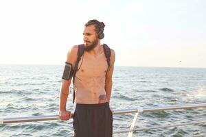 Young sporty bearded guy, rest after morning yoga at the seaside, listen favorite mix on headphones. photo