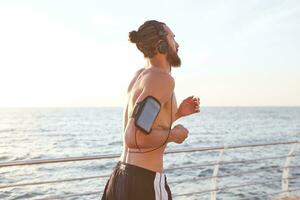 foto de joven hermoso deportivo barbado chico, escucha nuevo frio música en auriculares y corriendo a el playa. disfrutar el Mañana y puesta de sol.