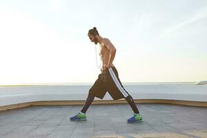 Photo of bearded young man with fit athletic body, does stretching exercises, listening music on headphones, has muscular body shape.