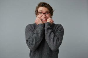 Portrait of young scared man with glasses wears in gray sweater, stands over gray background and bites nails. photo