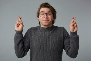 Photo of young handsome cheerful man with glasses wears in gray sweater, stands over gray background, crossed fingers and hopes for luck.