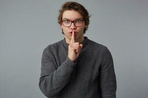 Make silence please Young man with glasses in gray sweater, shows silence sign, stands over gray background and keeps fore finger on lips, tries to keep conspiracy. photo