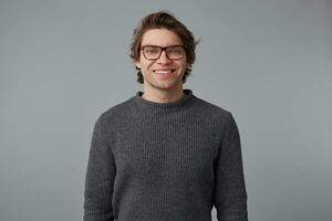 retrato de joven alegre hermoso hombre con lentes usa en gris suéter, soportes terminado gris antecedentes y en general sonrisas foto