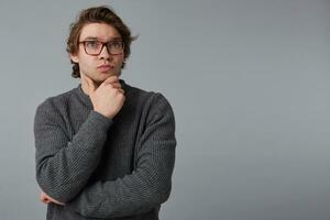 Portrait of young thinking man with glasses wears in gray sweater, stands over gray background with copy space on the right side, touches chin and looks away. photo