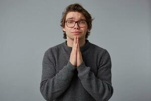 Young man with glasses wears in gray sweater, stands over gray background and looks at the camera, has sorrorful expression, keeps palms in praying gesture. photo