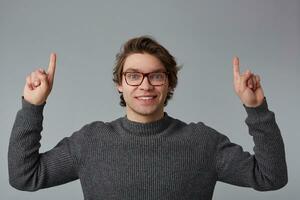 Portrait of young cheerful handsome man with glasses wears in gray sweater, stands over gray background and broadly smiles, points fingers up at a copy space over his head. photo