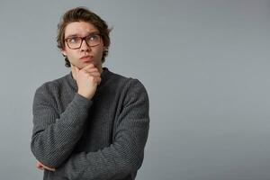 Portrait of young pensive man with glasses wears in gray sweater, stands over gray background with copy space on the right side, touches chin and looks up. photo