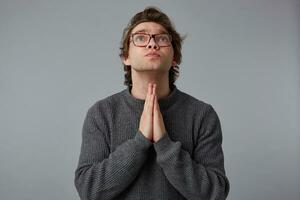 Portrait of young man with glasses wears in gray sweater, stands over gray background and looks up, has sorrorful expression, keeps palms in praying gesture, believes in something good. photo