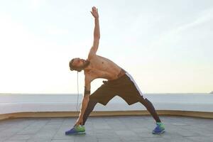 Young bearded jogger with fit athletic body, leans to feet, does stretching exercises, listening music on headphones, has muscular body shape. photo