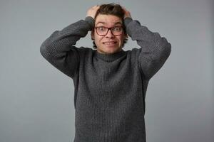 Portrait of young shocked attractive man with glasses wears in gray sweater, stands over gray background and lought, can't hold back emotions, holds hes head, look at the camera with wide open eyes. photo