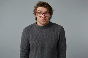 Portrait of young confused man with glasses wears in gray sweater, stands over gray background and looks at the camera with surprised expression. photo