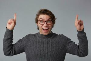 Portrait of young happy amazed man with glasses wears in gray sweater, stands over gray background with surprised expression, points fingers up at a copy space over his head. photo