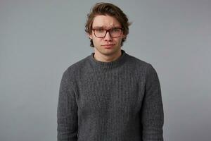 Photo of young sad handsome man with glasses wears in gray sweater, stands over gray background and looks unhappy.