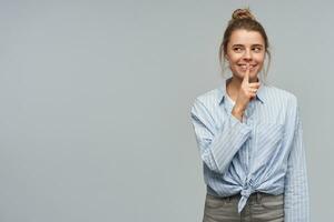 Young lady, pretty woman with blond hair bun. Wearing blue striped knotted shirt. Showing silence sign and playfully smiling. Watching to the left at copy space, isolated over grey background photo