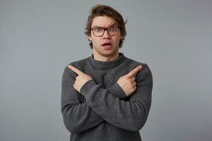 Portrait of young confused wondered man with glasses wears in gray sweater, stands over gray background, shows fingers in different directions. photo