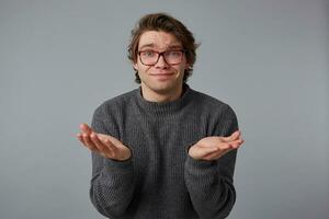 Photo of young pleaseful man with glasses wears in gray sweater, stands over gray background, and spreads his palms to the side.