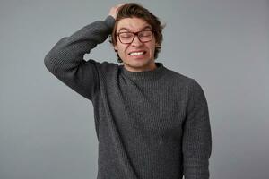 Portrait of young nervous man with glasses wears in gray sweater, stands over gray background and holds his head, feels severe migraine and pain. photo