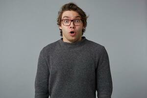 Portrait of young shocked man with glasses wears in gray sweater with wide open mouth and eyes, stands over gray background with surprised expression. photo