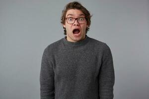 Portrait of young shocked guy with glasses, stands over gray background with wide open mouth and eyes, with surprised expression, looks scared. photo